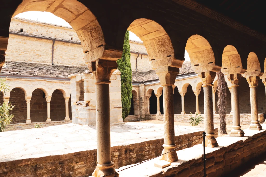 Cloister of the former cathedral of Roda de Isábena, with inscriptions in the arches and a cypress tree in the courtyard.
