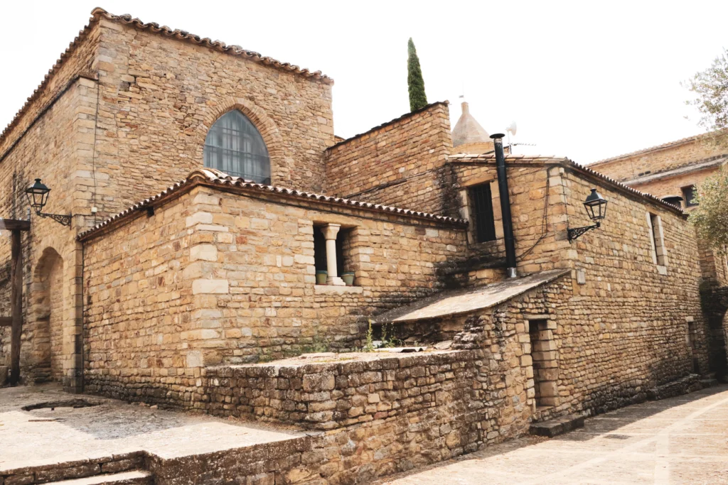 Exterior view of medieval stone buildings belonging to the Roda de Isábena cathedral complex, featuring tiered tiled roofs, a Gothic arched window, wall-mounted lanterns, and a cypress tree rising above the masonry walls.