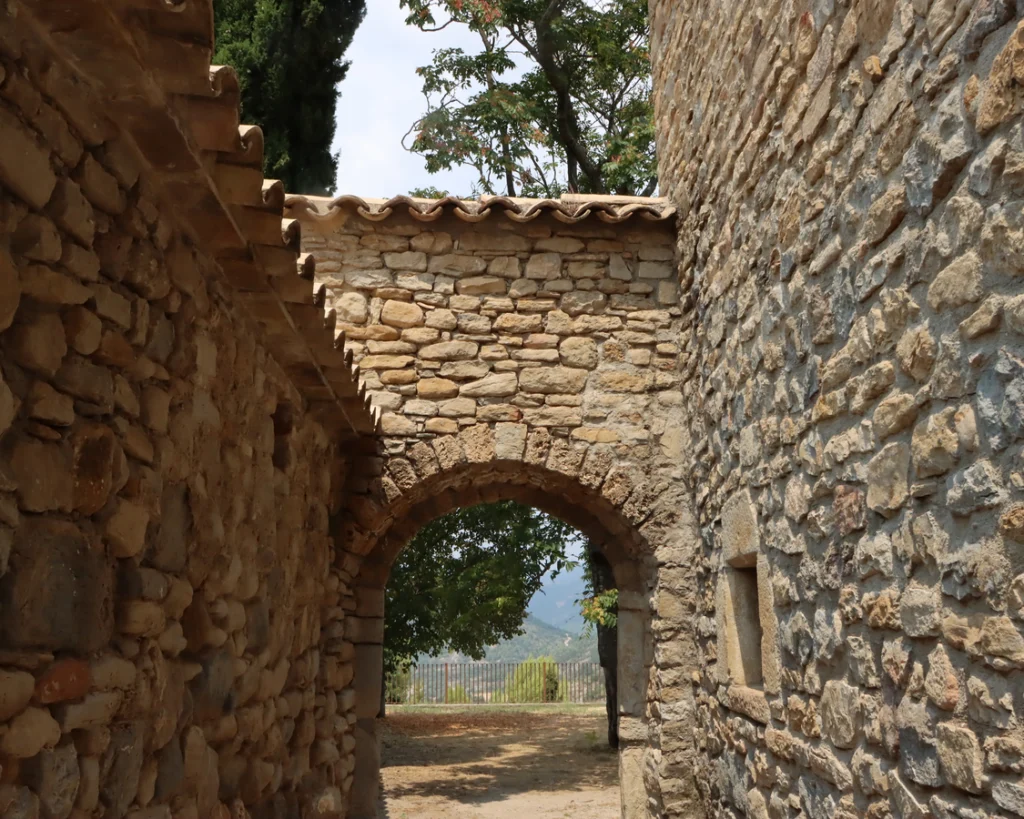 View through a rustic stone archway built between historic masonry walls in Roda de Isábena, looking out towards green trees and distant mountains.